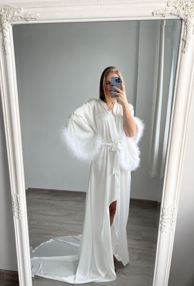 a woman taking a selfie in front of a mirror wearing a white robe and feathered shawl