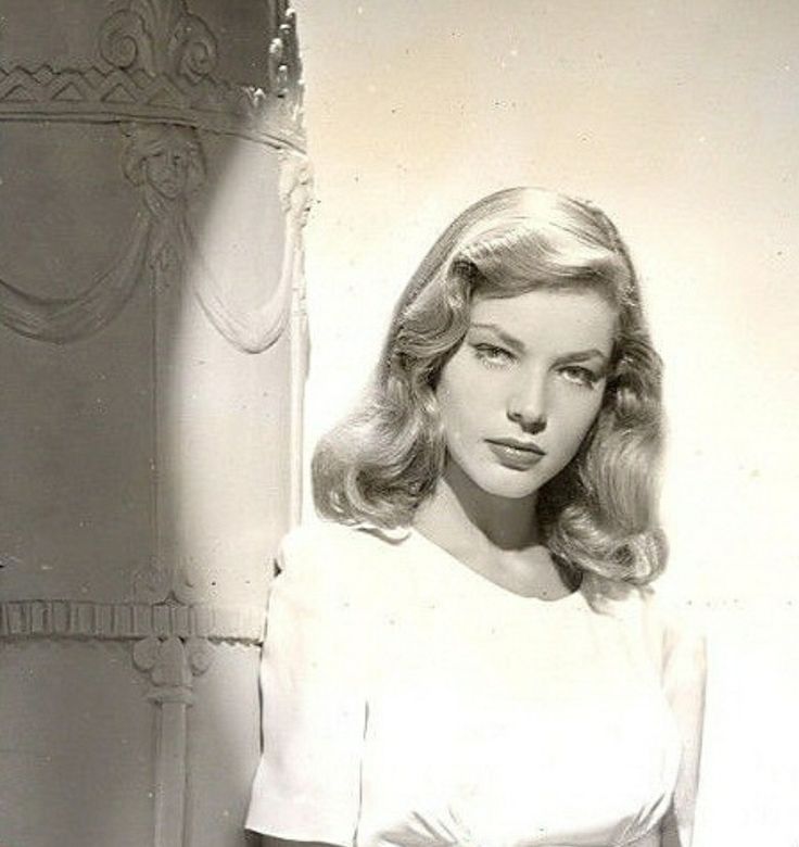 an old black and white photo of a woman with long hair wearing a dress in front of a wall