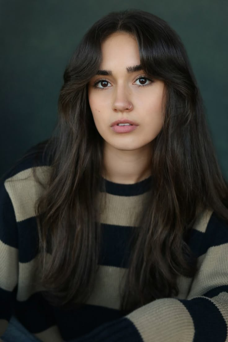 a woman with long dark hair wearing a striped sweater and looking at the camera while sitting in front of a green background