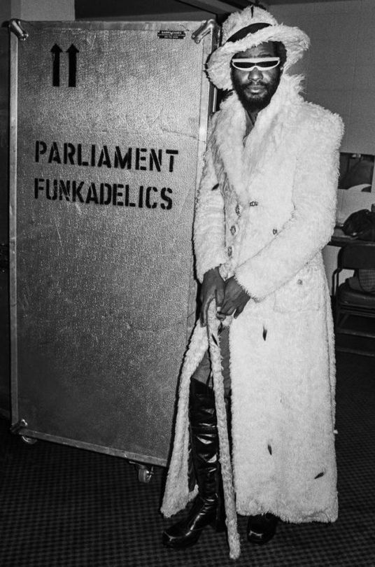 a man in a white coat and hat standing next to a metal box with the words parliament funkyadelics written on it