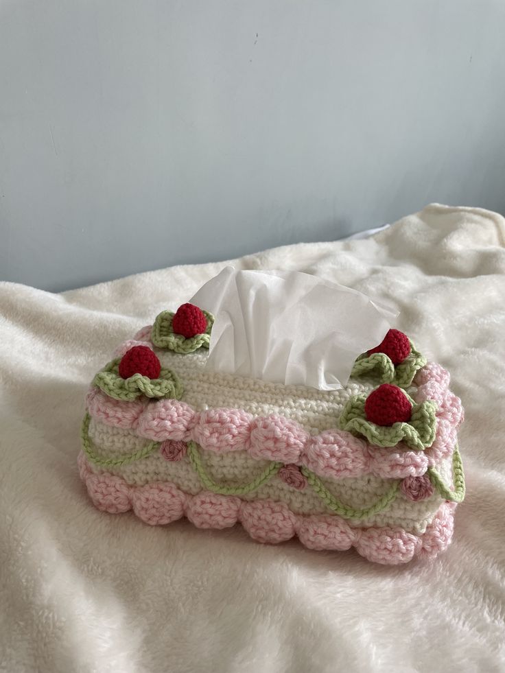 a pink and green crocheted tissue box on a white blanket with cherries