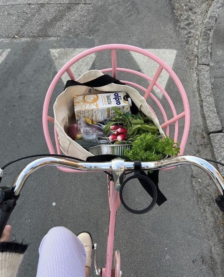 a pink bike with a basket full of food on the front handlebars and someone's feet