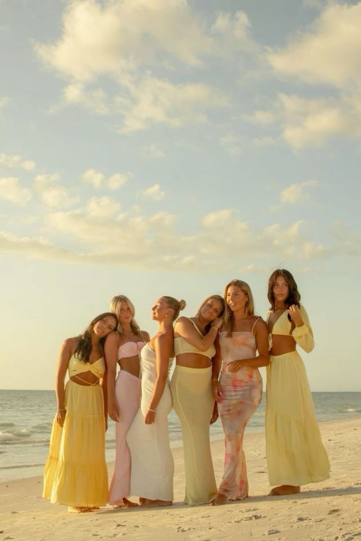 four women in dresses standing on the beach with their arms around each other's shoulders