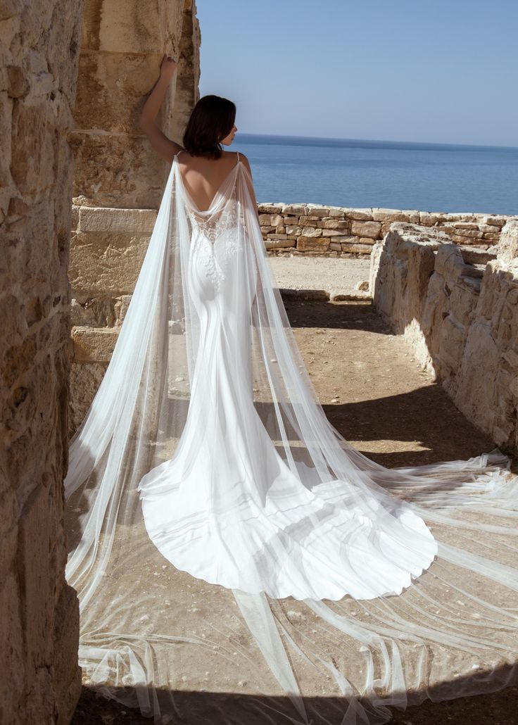 a woman in a wedding dress is standing near the ocean with her veil flying over her head