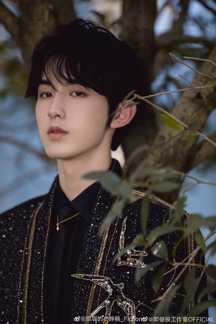 a young man standing next to a tree wearing a black shirt and gold trimmings