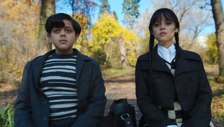 two people are sitting on a bench in the woods, one is wearing a coat