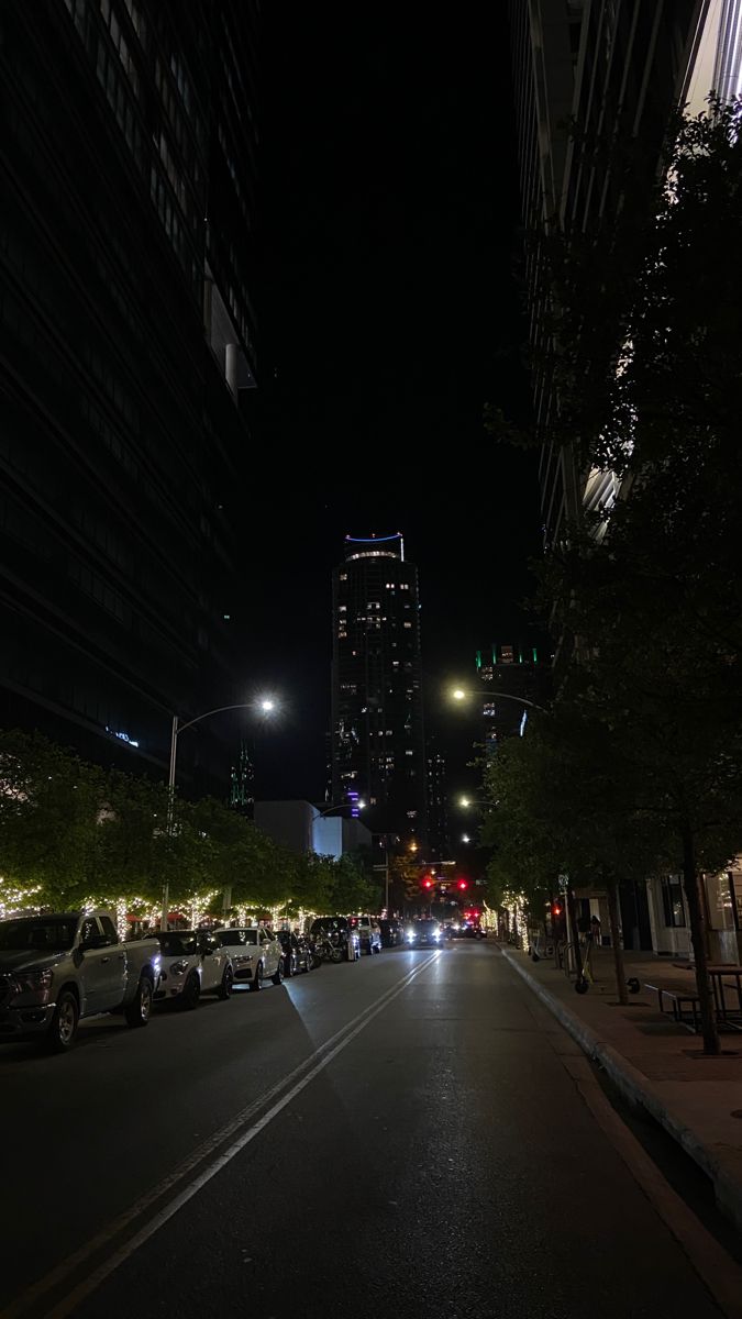 a city street at night with cars parked on both sides and tall buildings in the background