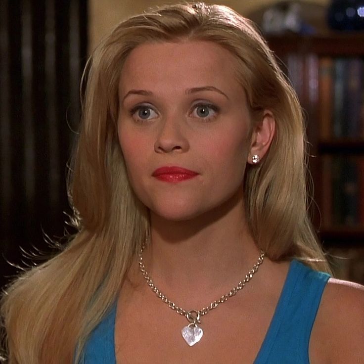 a blonde woman wearing a blue top with a heart shaped pendant on her neck, looking at the camera