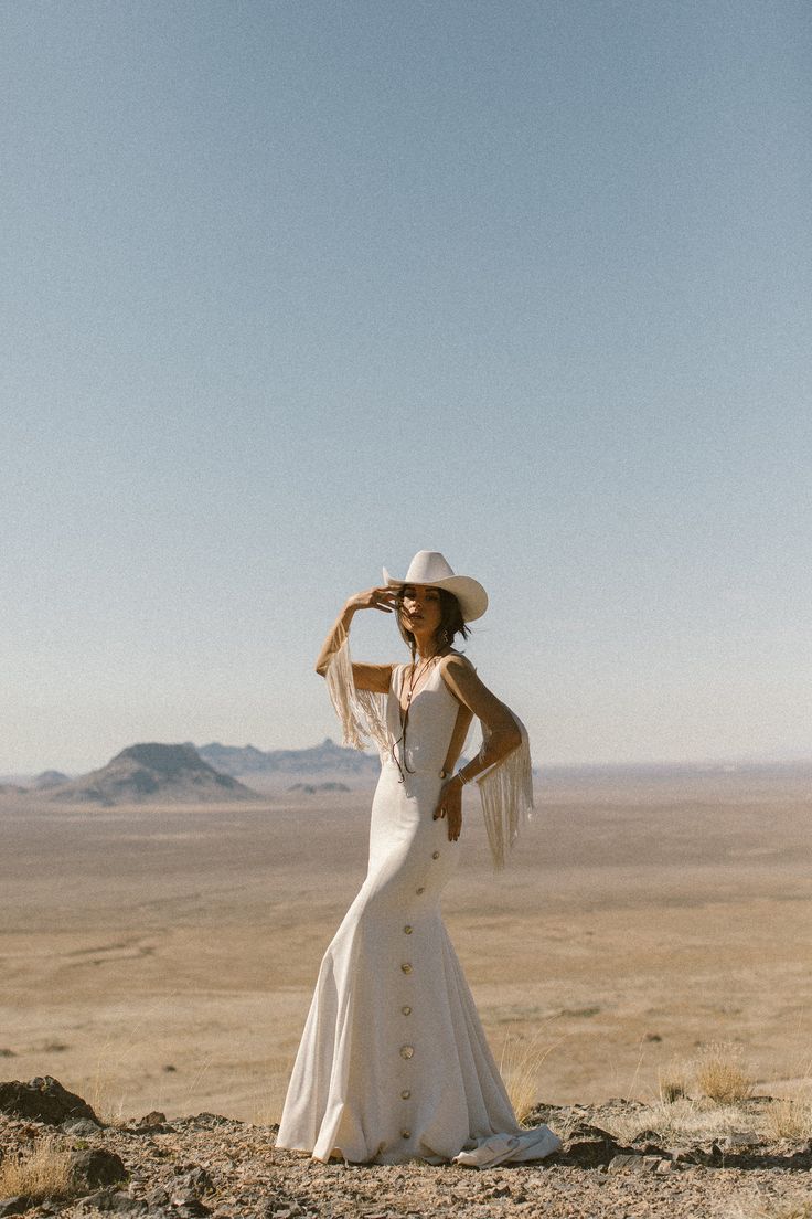 a woman in a white dress and hat standing on top of a dirt hill with mountains in the background