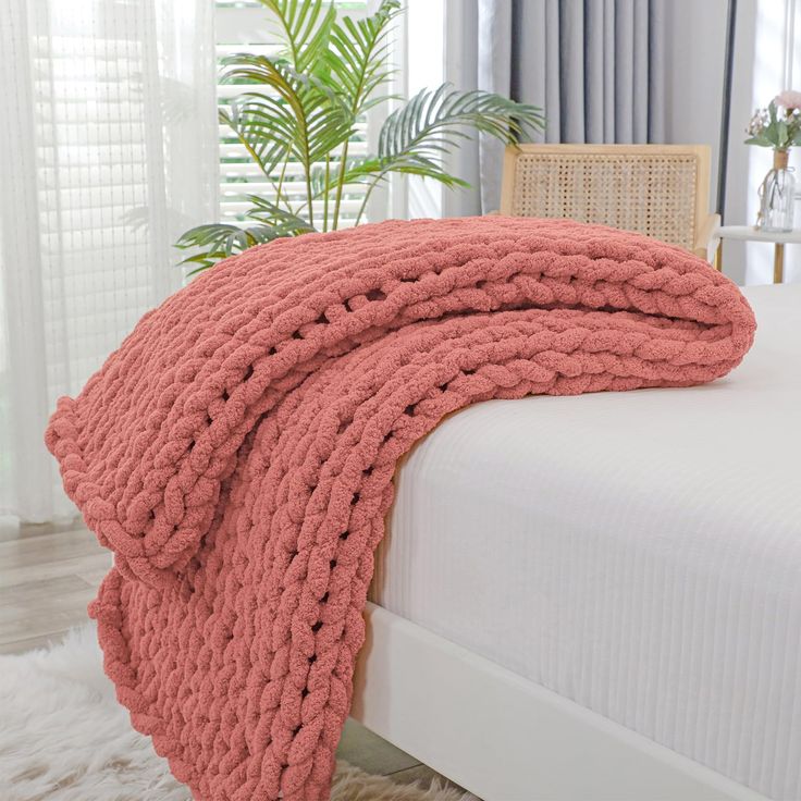 a pink knitted blanket on top of a white bed in a room with windows