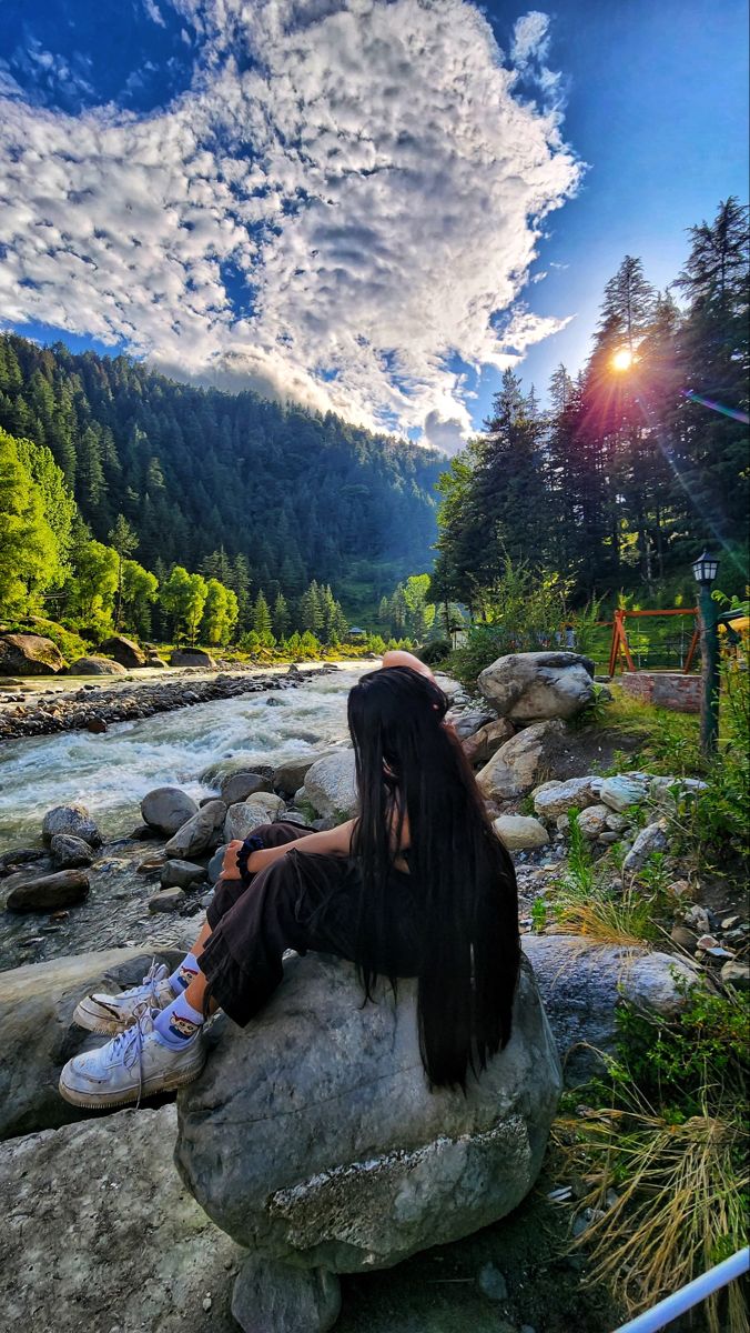 a woman sitting on top of a rock next to a river under a cloudy sky