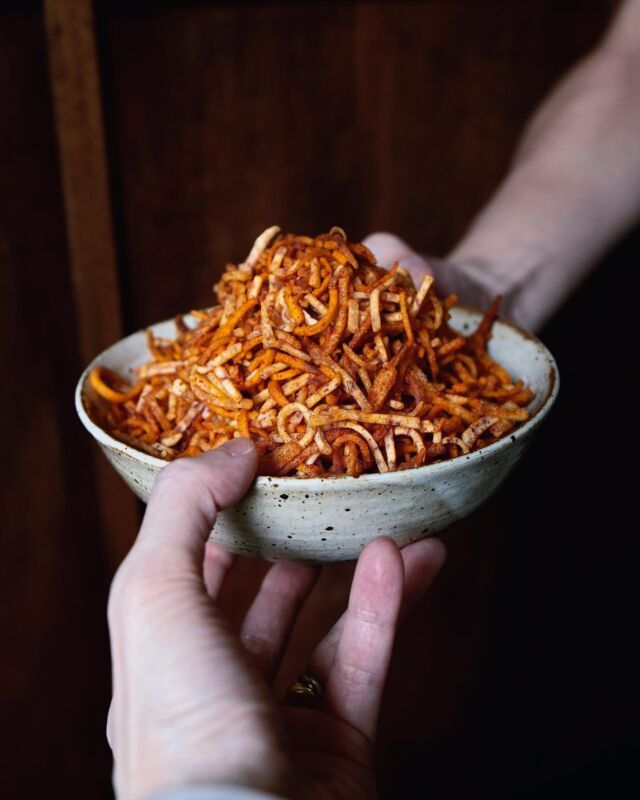 a person is holding a bowl full of fried noodles in their hands, with the other hand reaching for it