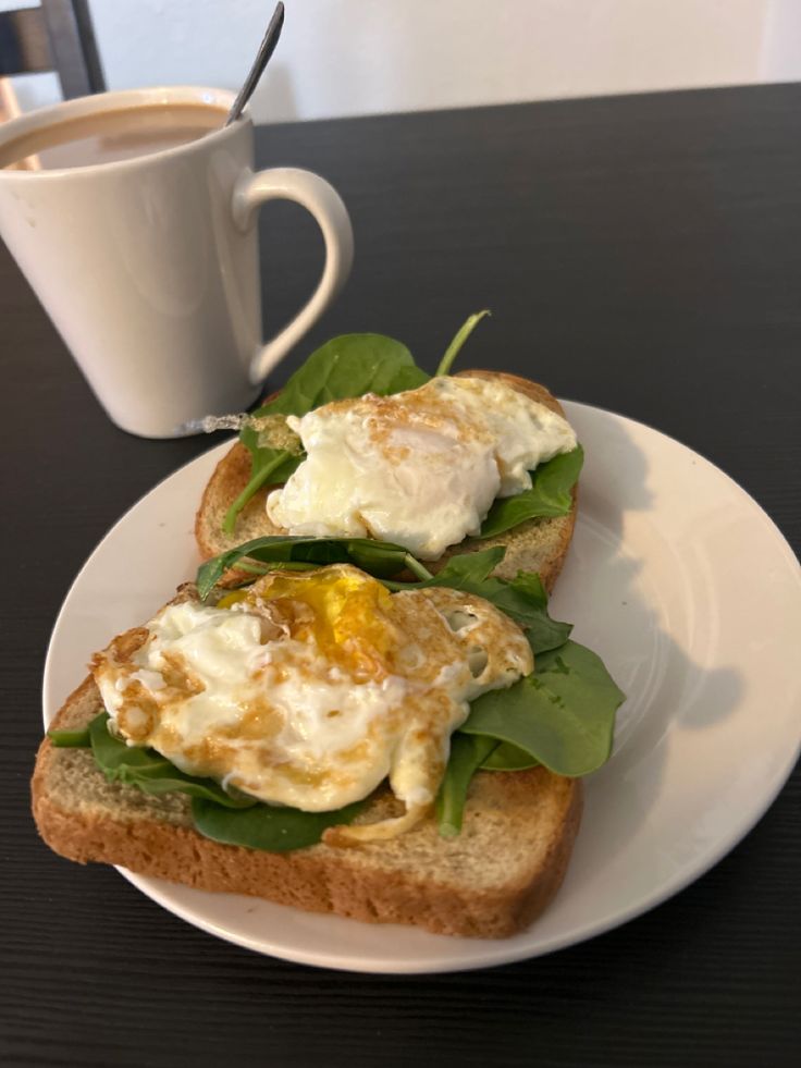 an open face sandwich with eggs and spinach sits on a plate next to a cup of coffee