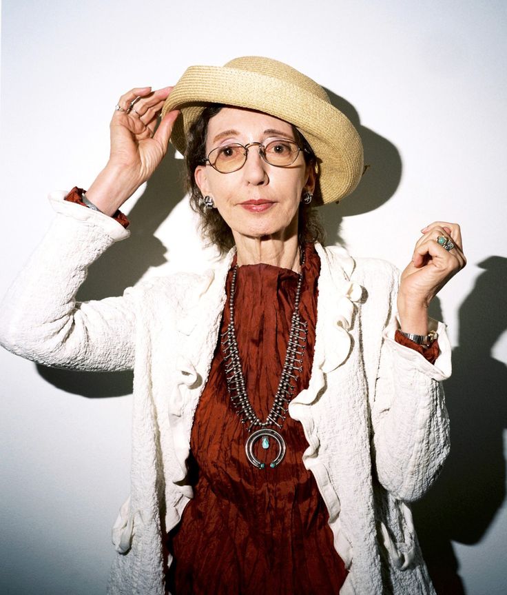 an older woman wearing glasses and a hat poses for a photo in front of a white wall