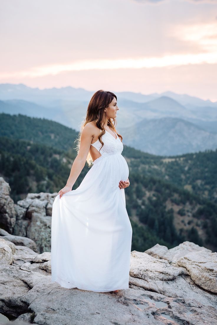 a pregnant woman in a white dress standing on top of a rocky hill with mountains in the background