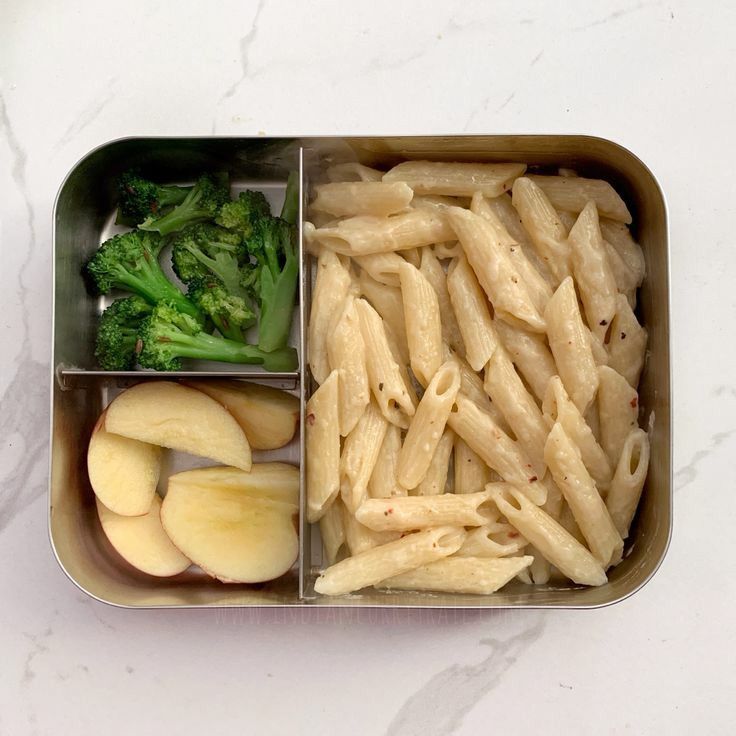 a metal container filled with pasta, broccoli and apple slices on top of a white counter