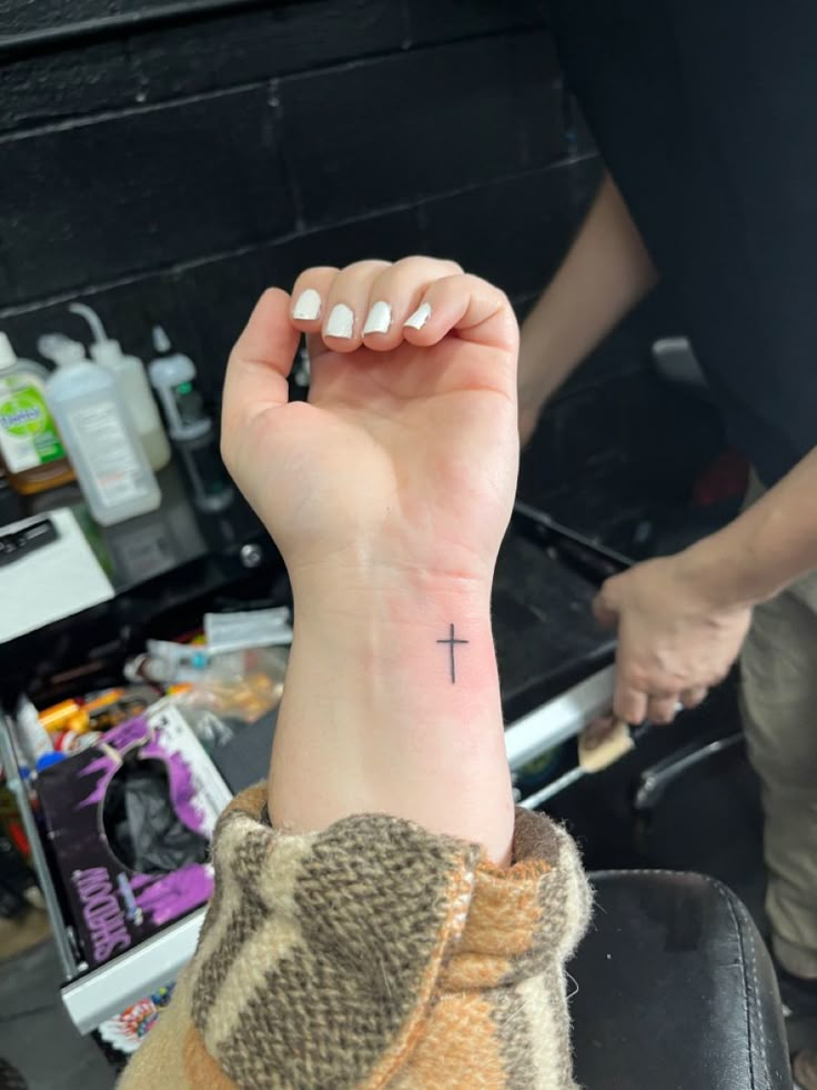 a person with a small cross tattoo on their wrist and hand, sitting in a chair