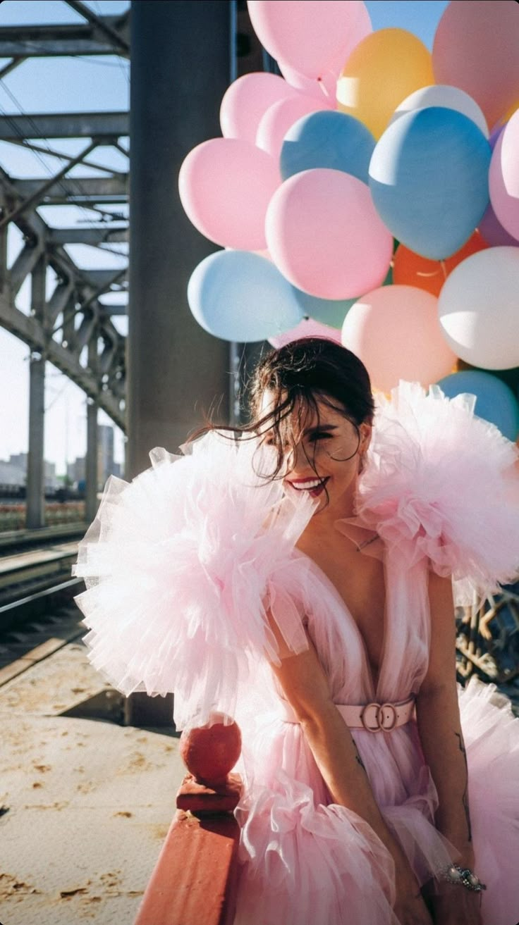 a woman in a pink dress sitting on a bench next to balloons and a train track