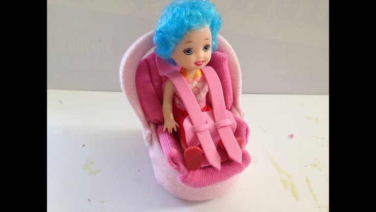 a doll sitting in a pink car seat with blue hair and wearing a pink sweater