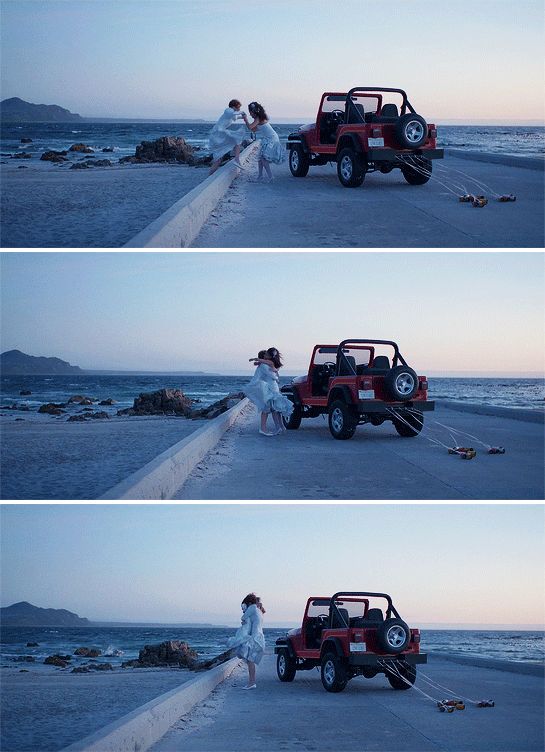 two people standing next to a red jeep on the beach at dusk, and another person in white