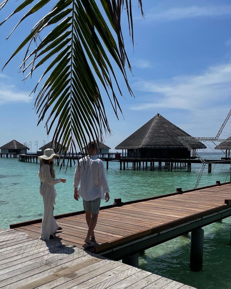 a man and woman walking on a dock towards the water with thatched huts in the background