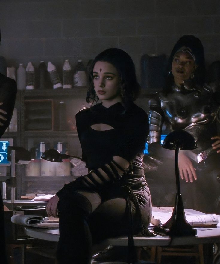 three women dressed in futuristic garb sitting at a table with an iron man behind them