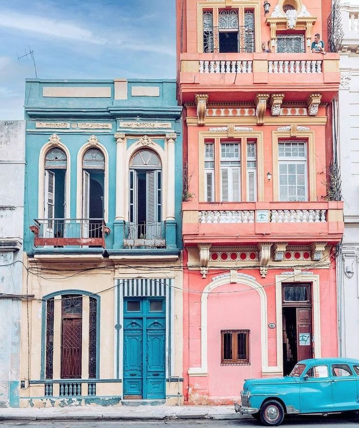 an old blue car parked in front of two multicolored buildings