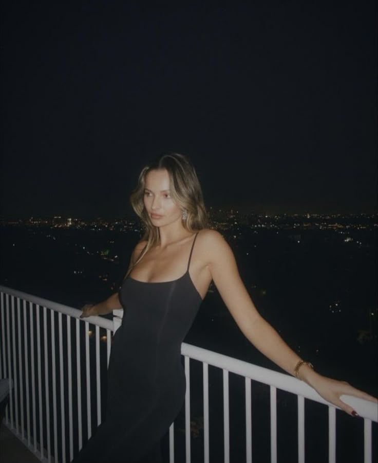 a woman in a black dress standing on a balcony at night with city lights behind her