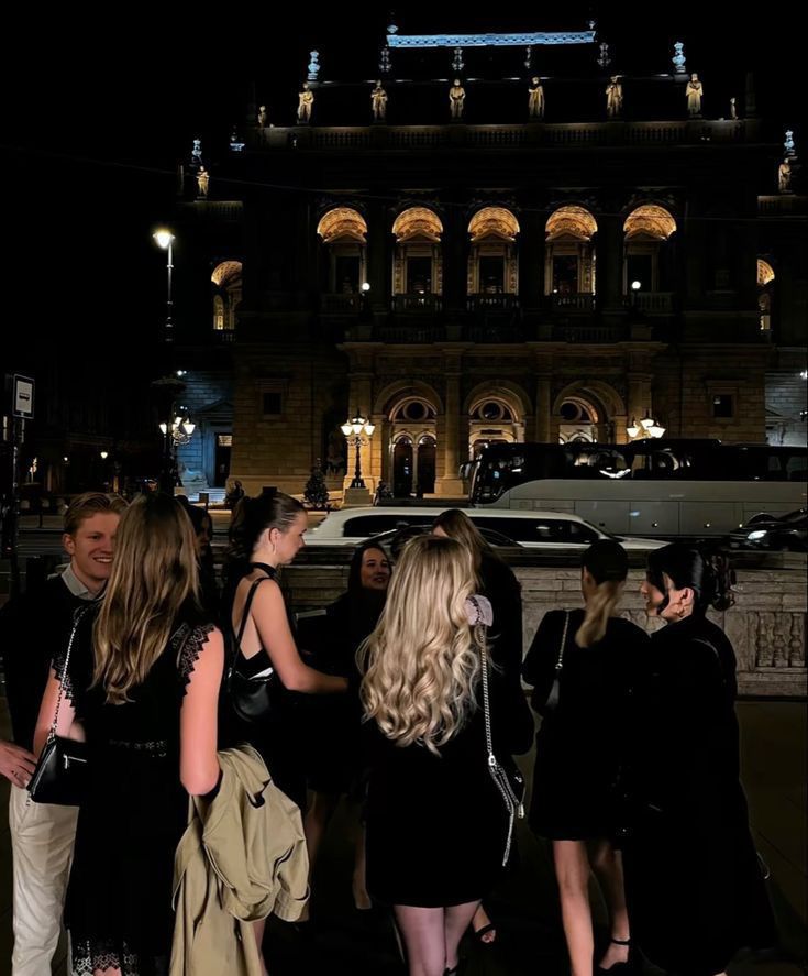 a group of people standing in front of a building at night with cars parked on the street