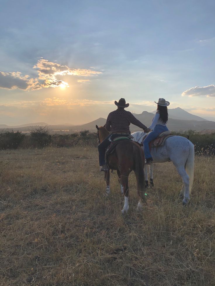 two people are riding horses in an open field with mountains in the background at sunset