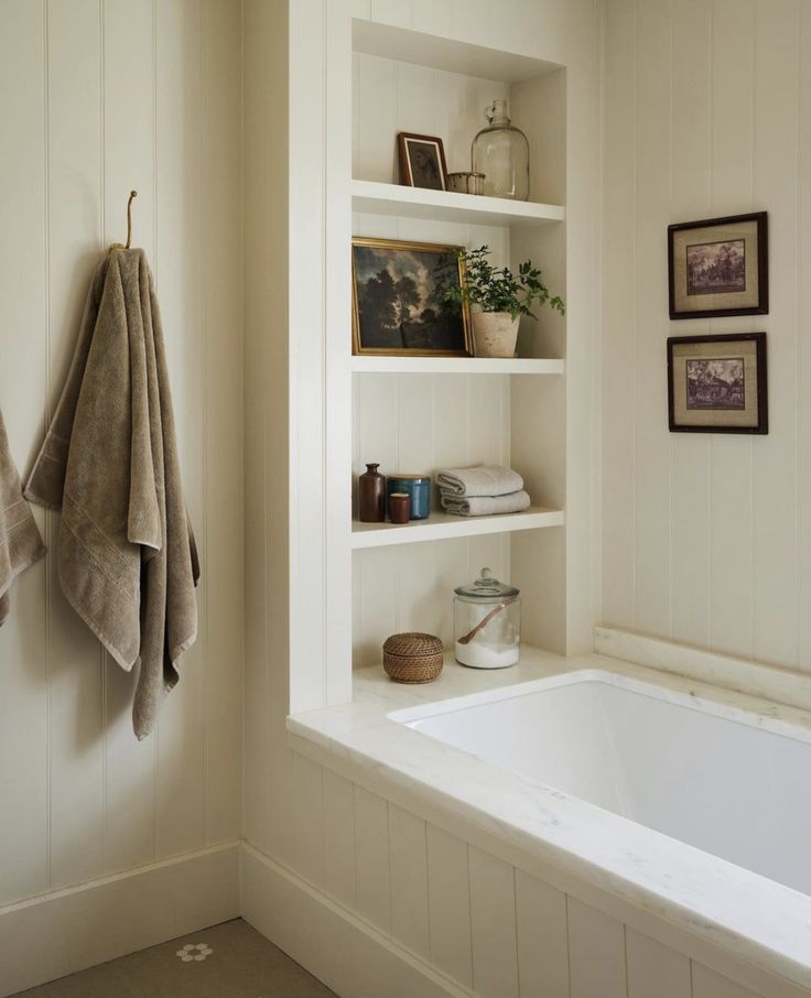 a bath tub sitting under a window next to a white shelf filled with books and pictures