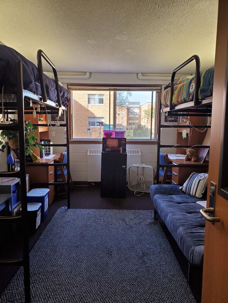 a dorm room with bunk beds and desks in the corner, next to a window