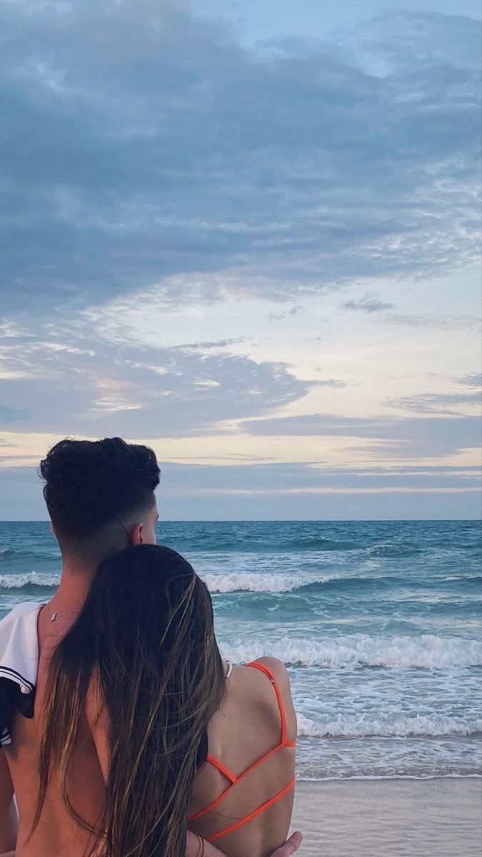 a man and woman sitting on top of each other at the beach looking out into the ocean