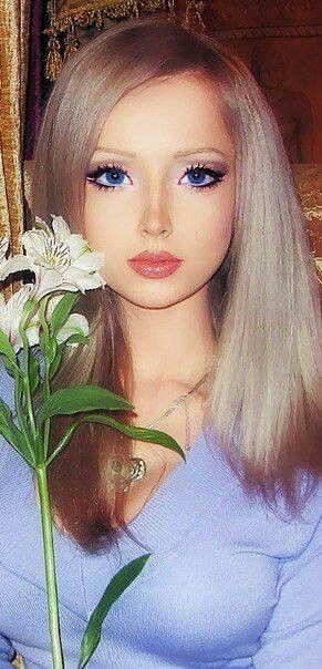 a doll with long blonde hair and blue eyes holds a flower in front of her face