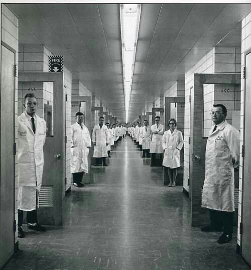 black and white photograph of men in lab coats lined up at the end of a hallway