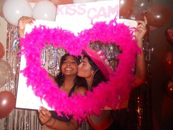 two women holding up a heart shaped sign in front of balloons and confetti