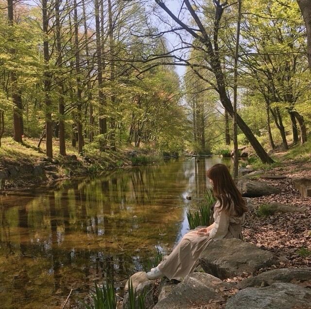 a woman sitting on top of a rock next to a river surrounded by trees and grass