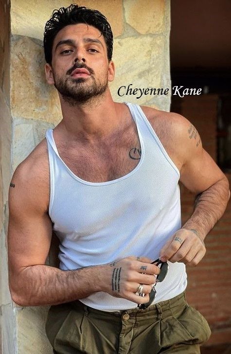 a man with tattoos standing next to a stone wall wearing green shorts and a white tank top