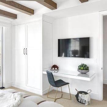 a bedroom with white walls and wood beams, a desk in the corner, and a flat screen tv mounted to the wall