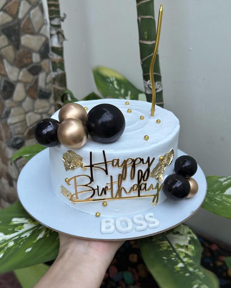 a person holding a birthday cake with gold and black decorations on it, in front of a plant