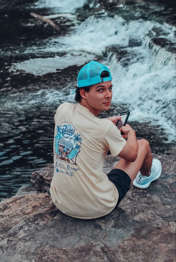 a young man sitting on top of a rock next to a river holding a cell phone