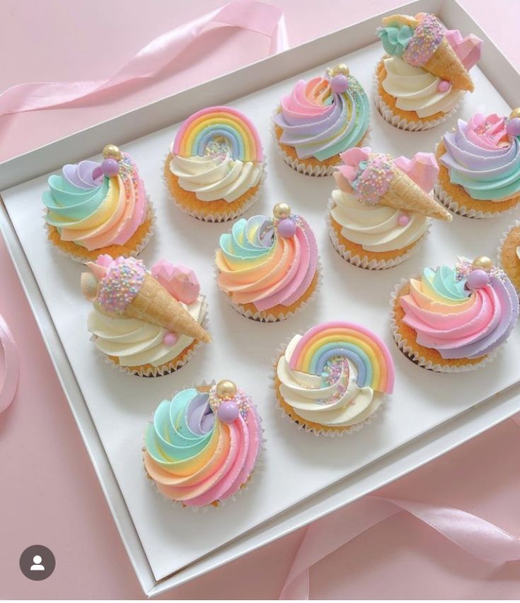 cupcakes decorated with rainbow icing and ice cream on a white tray next to pink ribbon