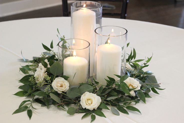 candles and flowers are arranged on a white table cloth with greenery around it for an elegant centerpiece