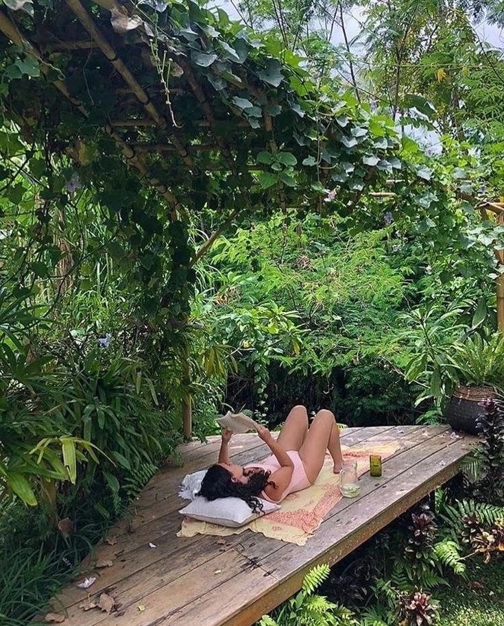 a woman laying on top of a wooden bench in the middle of a lush green forest