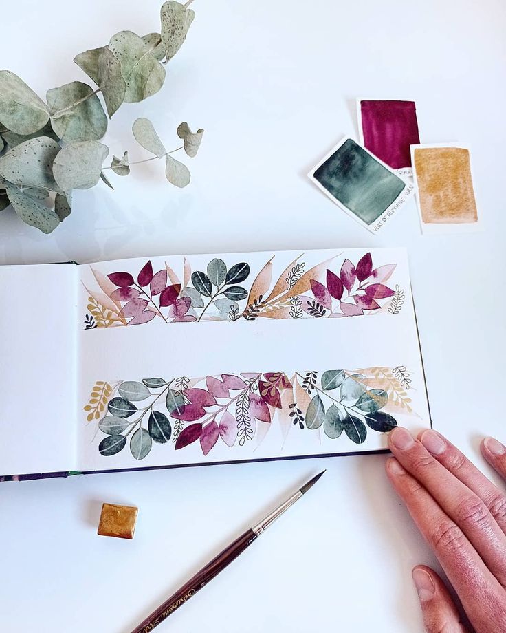 a hand is holding a pencil near some watercolors and other items on a table