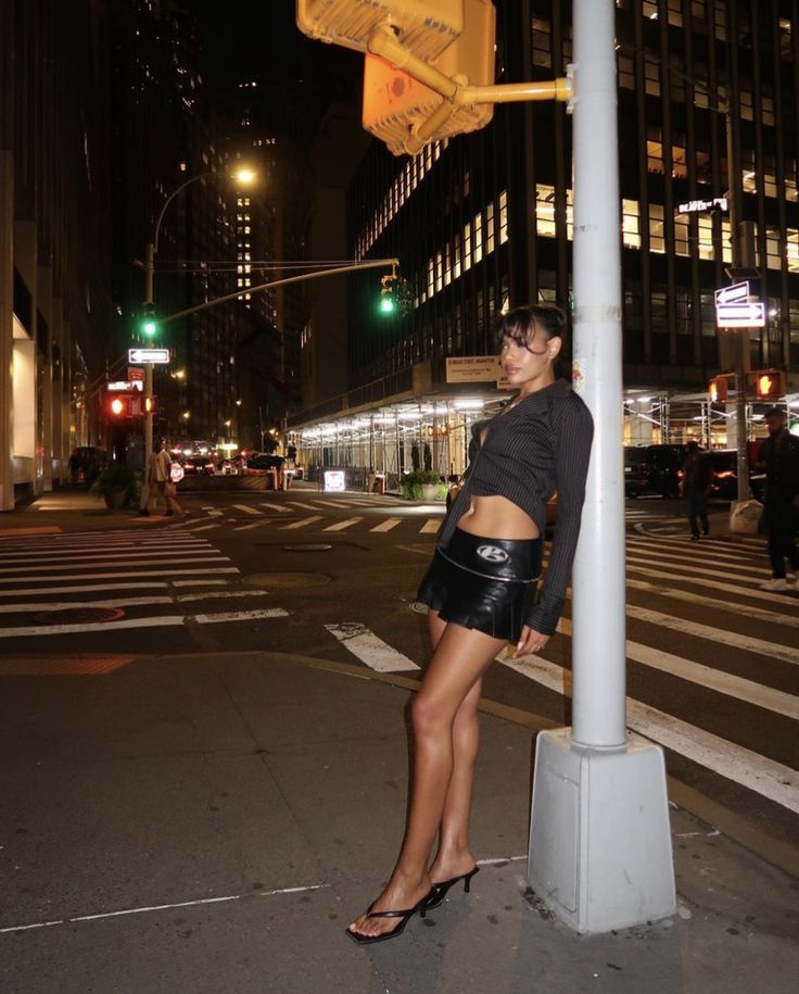 a woman leaning on a pole in the middle of an intersection at night with buildings and street lights behind her