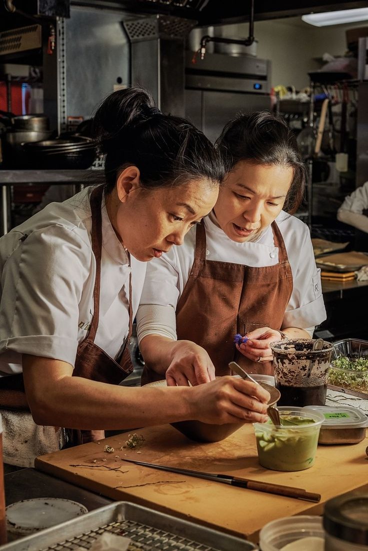 two women in aprons are preparing food on a cutting board with chopsticks