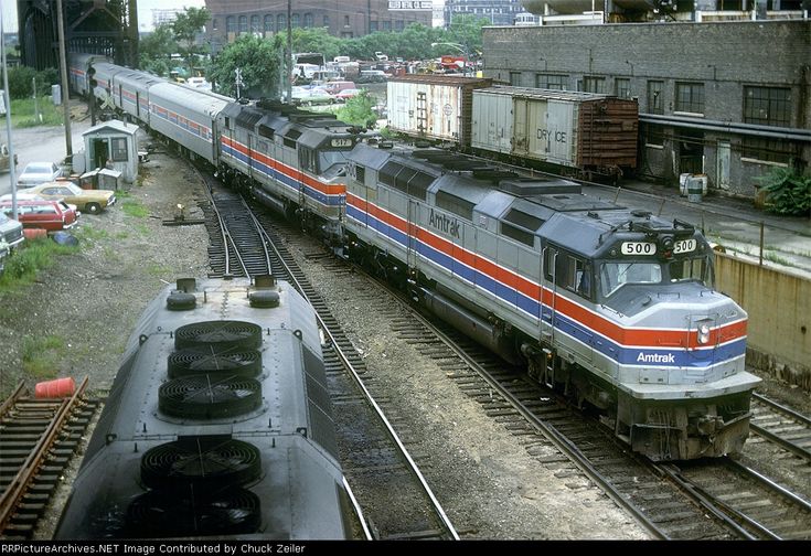 two trains passing each other on train tracks in an industrial area with buildings and trees