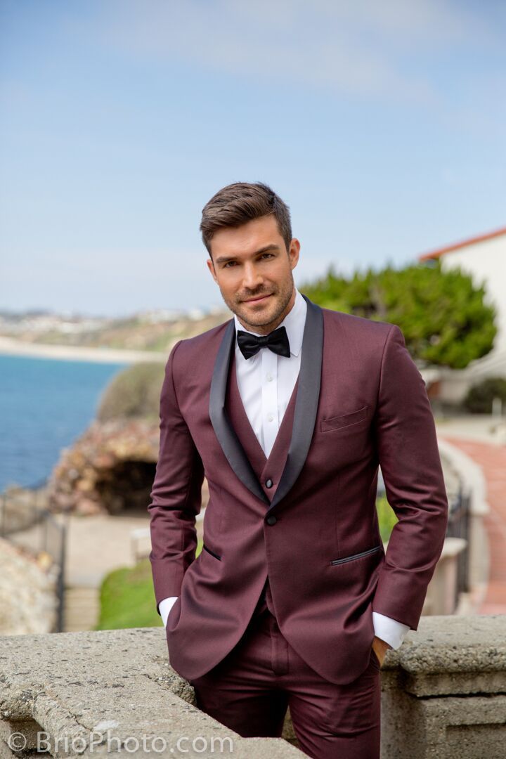 a man in a tuxedo standing next to a wall near the ocean with his hands on his hips