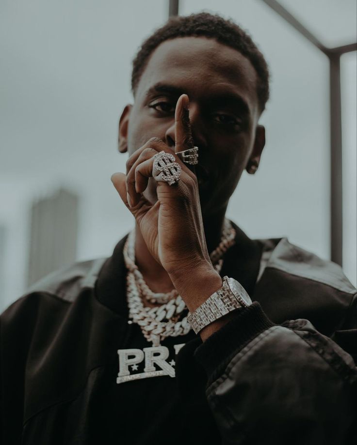 a man holding up his finger to the camera while wearing a black jacket and necklace